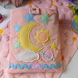 May include: A pink corduroy book cover featuring felt celestial appliques including a large yellow crescent moon, a sun, stars, a cloud, and a planet. A blue wavy trim runs along the bottom edge with a label reading Songbird Market Co.