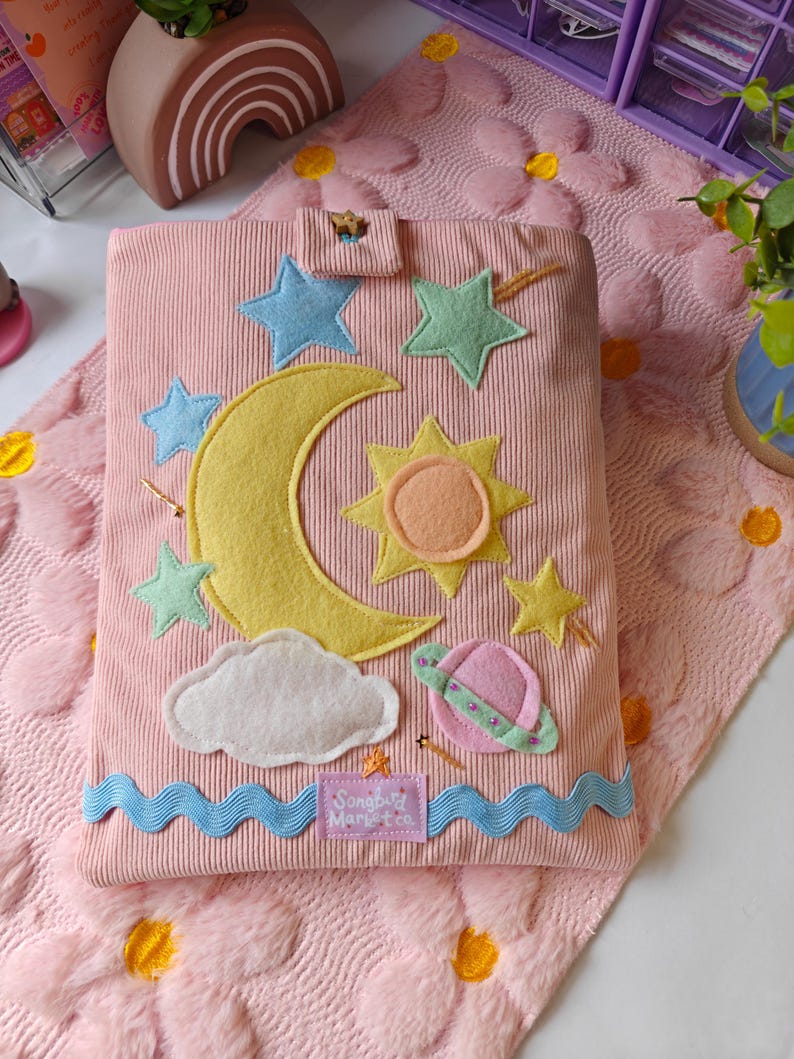 May include: A pink corduroy book cover featuring felt celestial appliques including a large yellow crescent moon, a sun, stars, a cloud, and a planet. A blue wavy trim runs along the bottom edge with a label reading Songbird Market Co.