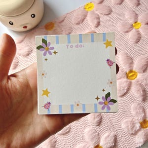 May include: A square notepad with a floral design and the words "To do:" printed at the top. The notepad has a light blue striped border and is decorated with purple flowers, stars, and ladybugs. The notepad is held in a hand.