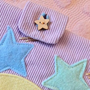 May include: A lavender corduroy fabric pouch featuring a wooden star-shaped button closure. The pouch is decorated with felt appliques including a blue star, a mint green star, a yellow crescent moon, and a peach sun with yellow rays.