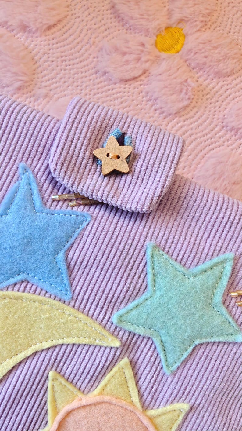 May include: A lavender corduroy fabric pouch featuring a wooden star-shaped button closure. The pouch is decorated with felt appliques including a blue star, a mint green star, a yellow crescent moon, and a peach sun with yellow rays.