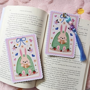 May include: Two illustrated bookmarks featuring a cute bunny wrapped in a green blanket, holding a cup. The bookmarks have a pink border with flowers, butterflies, and strawberries. A blue tassel is attached to one bookmark.