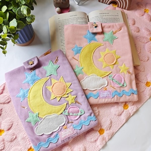 May include: Two rectangular corduroy book sleeves, one in lavender and one in light pink, featuring felt appliqu&eacute;s of yellow crescent moons, suns, stars, clouds, and planets. Each sleeve has a button closure and a small label reading Sunbird Market Co.