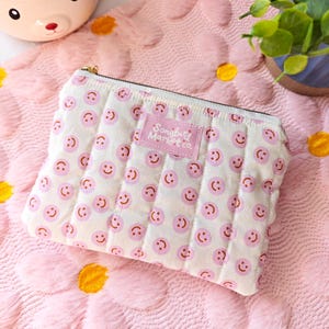 May include: A quilted cosmetic bag with a white background and a pattern of pink smiley faces. The bag has a gold zipper and a pink label that reads "Songbud Market co.". The bag is on a pink daisy-patterned surface.