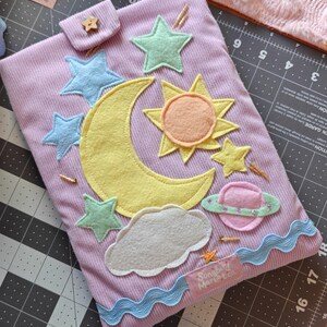 May include: A lavender corduroy pouch featuring felt appliqu&eacute;s of a yellow crescent moon, a sun, a pink planet with a green ring, a gray cloud, and several blue and green stars. A small wooden star button secures the flap, and a label reads Songbird Market co.