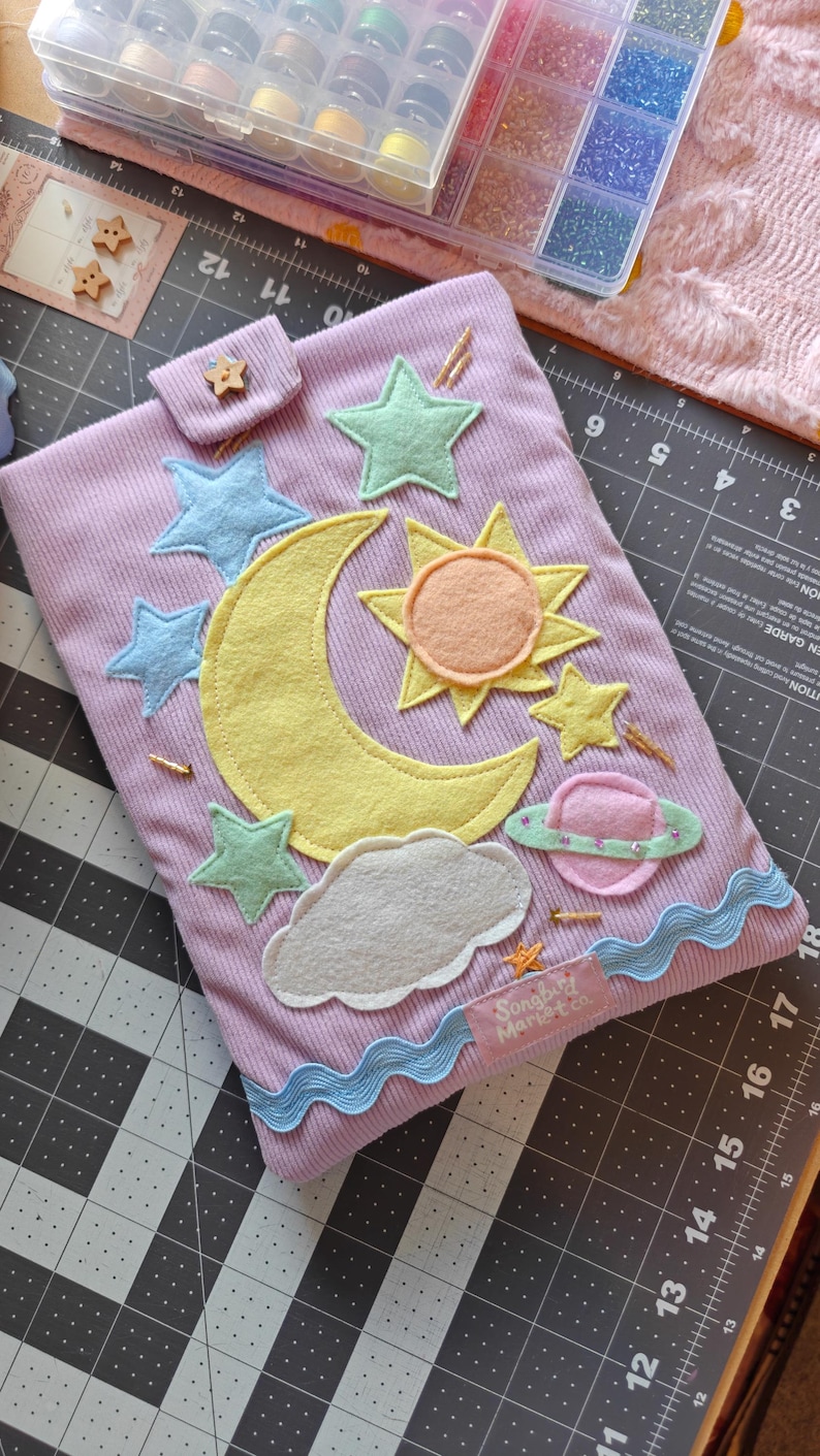 May include: A lavender corduroy pouch featuring felt appliqu&eacute;s of a yellow crescent moon, a sun, a pink planet with a green ring, a gray cloud, and several blue and green stars. A small wooden star button secures the flap, and a label reads Songbird Market co.