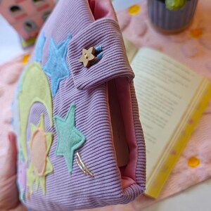May include: A lavender corduroy book sleeve featuring a light pink interior lining. The exterior is decorated with felt appliques of a yellow crescent moon, a sun, and light blue stars. A small wooden star-shaped button secures the flap closure.