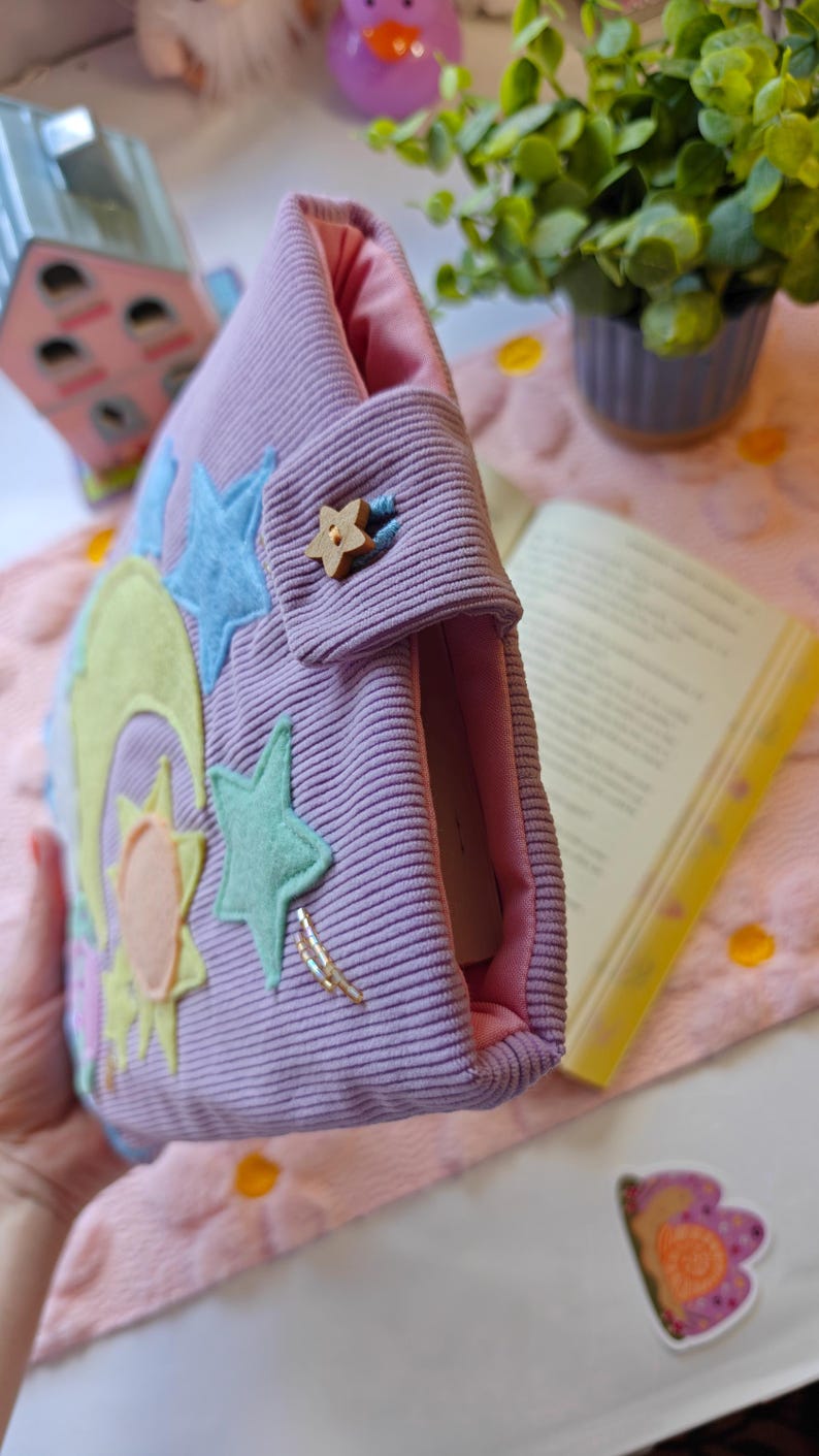 May include: A lavender corduroy book sleeve featuring a light pink interior lining. The exterior is decorated with felt appliques of a yellow crescent moon, a sun, and light blue stars. A small wooden star-shaped button secures the flap closure.