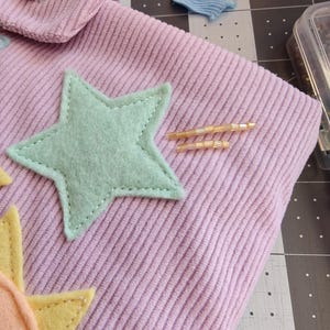 May include: A light purple corduroy fabric pouch featuring a mint green felt star appliqu&eacute;, a yellow felt sun shape, a small wooden star-shaped button, and two thin horizontal lines of light-colored glass beads stitched onto the surface.
