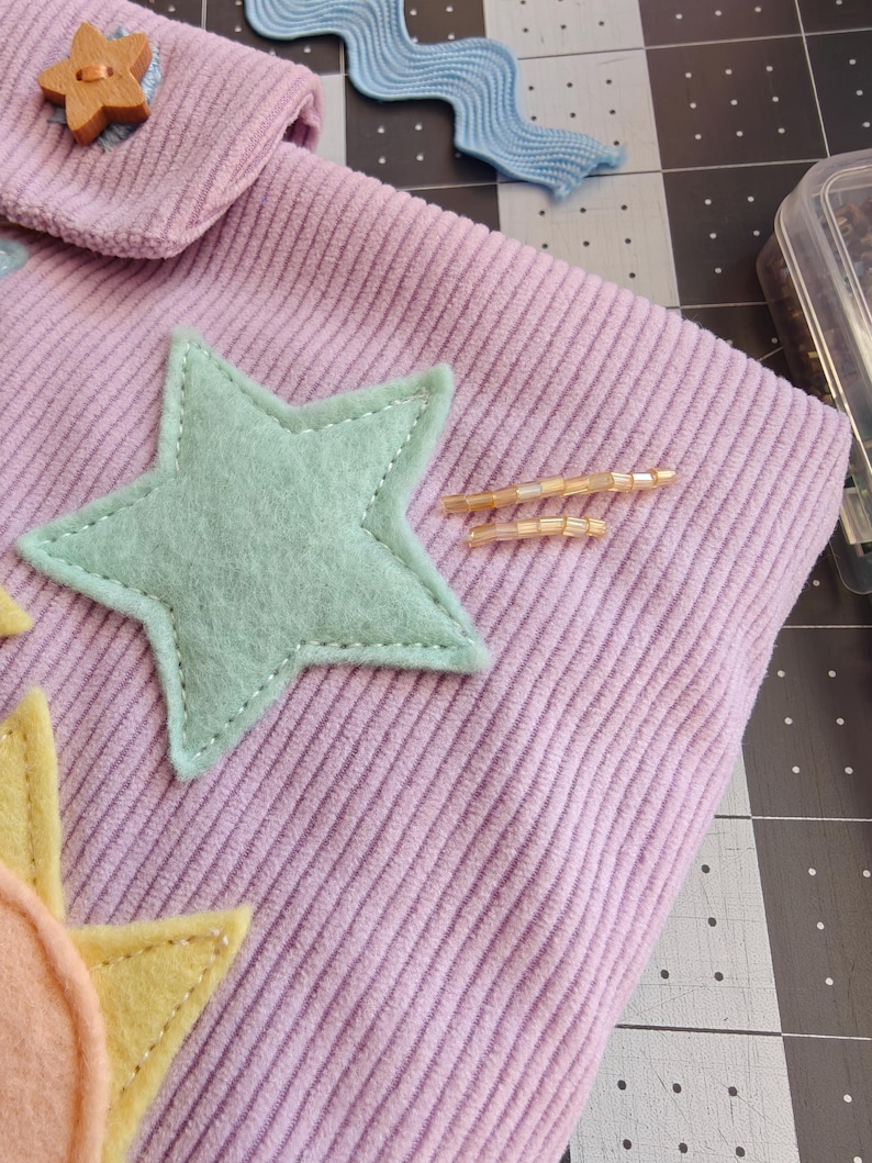 May include: A light purple corduroy fabric pouch featuring a mint green felt star appliqu&eacute;, a yellow felt sun shape, a small wooden star-shaped button, and two thin horizontal lines of light-colored glass beads stitched onto the surface.