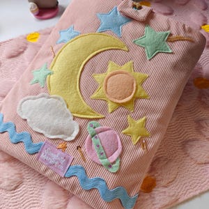 May include: A pink corduroy book cover featuring felt appliques of a yellow crescent moon, a sun, stars, a white cloud, and a pink planet with a green ring. A blue wavy ribbon trim and a purple fabric tag labeled Songbird Market Co. are attached.