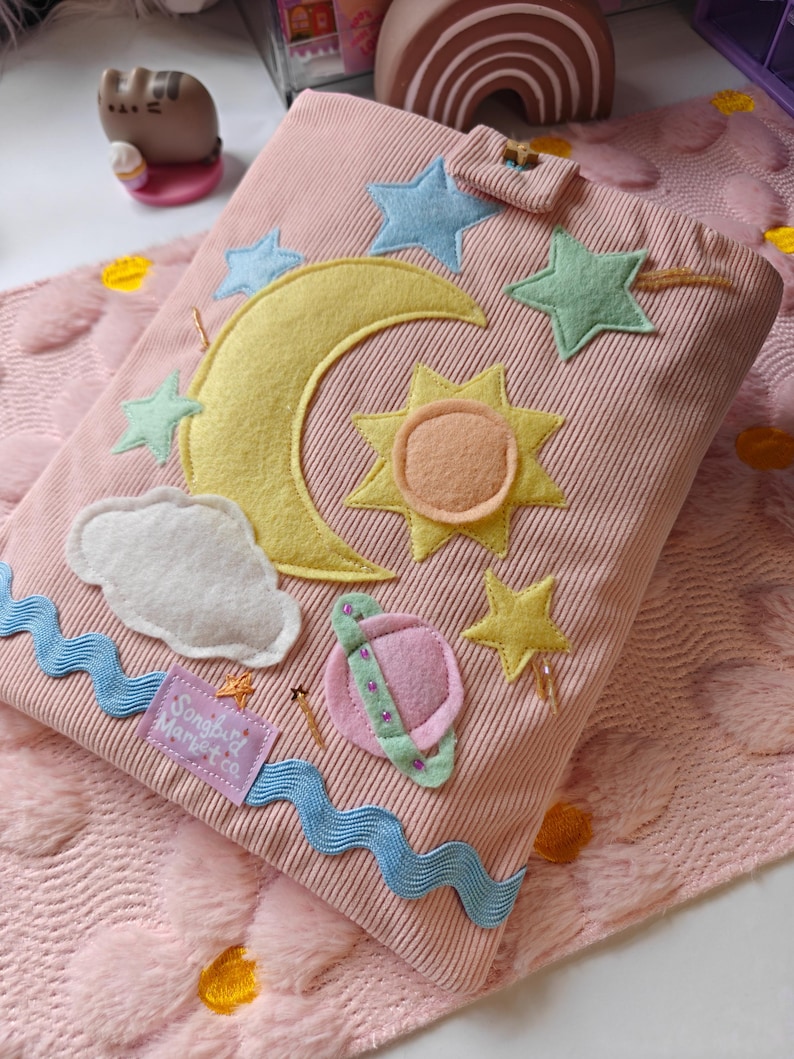 May include: A pink corduroy book cover featuring felt appliques of a yellow crescent moon, a sun, stars, a white cloud, and a pink planet with a green ring. A blue wavy ribbon trim and a purple fabric tag labeled Songbird Market Co. are attached.