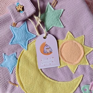 May include: A light purple corduroy pouch decorated with felt appliques of a large yellow crescent moon, a yellow sun, light blue and mint green stars, and a white cloud. A tag features a cartoon bird on a moon with the text Songbird Market co.