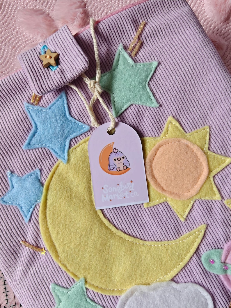 May include: A light purple corduroy pouch decorated with felt appliques of a large yellow crescent moon, a yellow sun, light blue and mint green stars, and a white cloud. A tag features a cartoon bird on a moon with the text Songbird Market co.