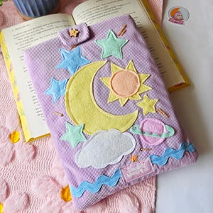 A handmade lavender corduroy book sleeve lies flat on a white surface, resting on a pink floral-patterned rug. The sleeve is decorated with colorful fabric appliqués of a large yellow crescent moon, a yellow and orange sun, a pink ringed planet, and several light blue and green stars. Small, hand-beaded gold details represent shimmering stars scattered across the design, and a wavy blue trim accents the bottom edge. A small wooden star-shaped button secures the top flap.