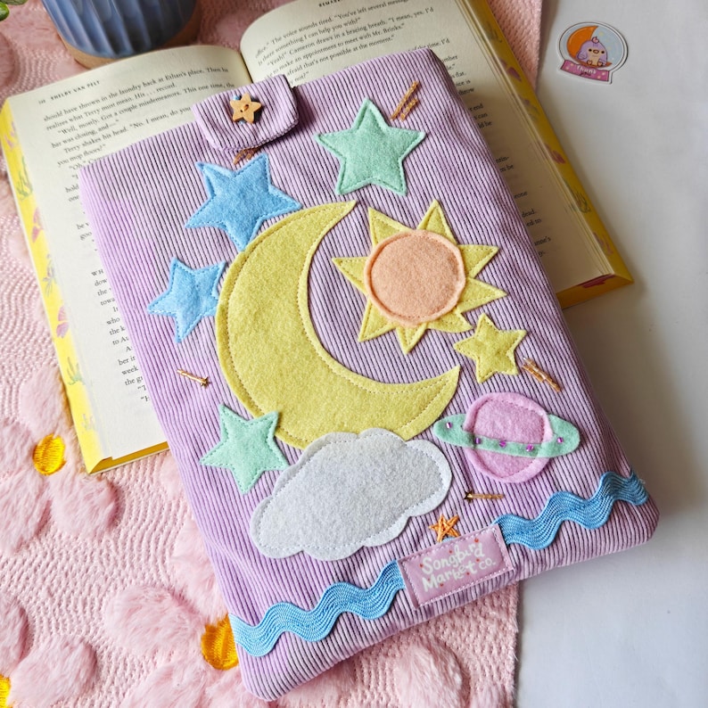 A handmade lavender corduroy book sleeve lies flat on a white surface, resting on a pink floral-patterned rug. The sleeve is decorated with colorful fabric appliqués of a large yellow crescent moon, a yellow and orange sun, a pink ringed planet, and several light blue and green stars. Small, hand-beaded gold details represent shimmering stars scattered across the design, and a wavy blue trim accents the bottom edge. A small wooden star-shaped button secures the top flap.