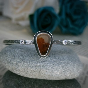 Puede incluir: Un brazalete de plata con una piedra de &aacute;gata en forma de l&aacute;grima engastada en un bisel de plata. La piedra de &aacute;gata tiene un patr&oacute;n marr&oacute;n y blanco. El brazalete tambi&eacute;n tiene dos peque&ntilde;as piedras transparentes colocadas a cada lado de la piedra de &aacute;gata.