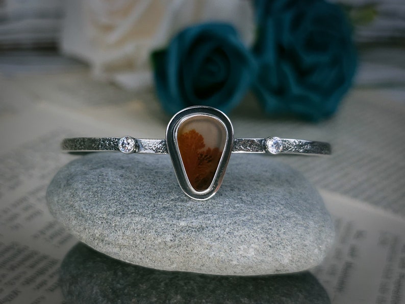 Puede incluir: Un brazalete de plata con una piedra de &aacute;gata en forma de l&aacute;grima engastada en un bisel de plata. La piedra de &aacute;gata tiene un patr&oacute;n marr&oacute;n y blanco. El brazalete tambi&eacute;n tiene dos peque&ntilde;as piedras transparentes colocadas a cada lado de la piedra de &aacute;gata.
