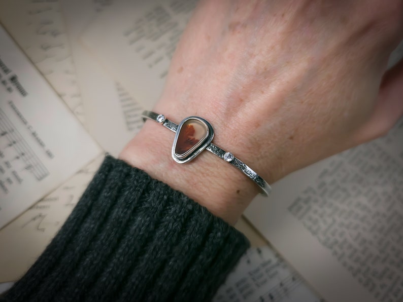 Puede incluir: Un brazalete de plata con una piedra de &aacute;gata en forma de l&aacute;grima engastada en un bisel de plata. El brazalete tiene tres peque&ntilde;os detalles de plata.