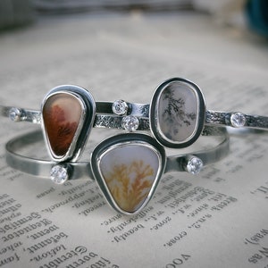 Puede incluir: Tres pulseras de plata con detalles de piedra de &aacute;gata. Cada pulsera presenta una piedra de &aacute;gata diferente con patrones y colores &uacute;nicos. Las piedras est&aacute;n engastadas en biseles de plata y las pulseras est&aacute;n adornadas con peque&ntilde;os cristales transparentes.