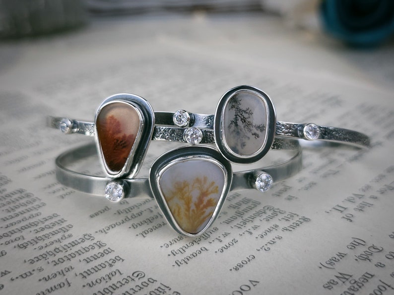 Puede incluir: Tres pulseras de plata con detalles de piedra de &aacute;gata. Cada pulsera presenta una piedra de &aacute;gata diferente con patrones y colores &uacute;nicos. Las piedras est&aacute;n engastadas en biseles de plata y las pulseras est&aacute;n adornadas con peque&ntilde;os cristales transparentes.