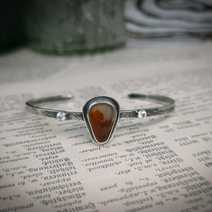 Puede incluir: Una pulsera de plata con una banda texturizada y un colgante en forma de l&aacute;grima con una piedra de &aacute;gata roja y marr&oacute;n. El colgante est&aacute; acentuado con dos peque&ntilde;as piedras de circonita c&uacute;bica transparentes.