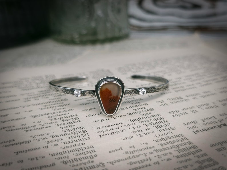 Puede incluir: Una pulsera de plata con una banda texturizada y un colgante en forma de l&aacute;grima con una piedra de &aacute;gata roja y marr&oacute;n. El colgante est&aacute; acentuado con dos peque&ntilde;as piedras de circonita c&uacute;bica transparentes.