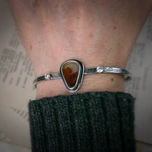 Puede incluir: Una pulsera de plata con una piedra de &aacute;gata en forma de l&aacute;grima engastada en un bisel de plata. La piedra tiene un patr&oacute;n marr&oacute;n y blanco. La pulsera tiene una peque&ntilde;a perla de plata a cada lado de la piedra.