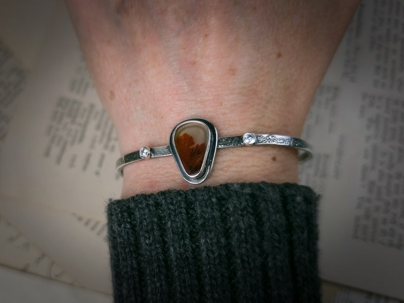 Puede incluir: Una pulsera de plata con una piedra de &aacute;gata en forma de l&aacute;grima engastada en un bisel de plata. La piedra tiene un patr&oacute;n marr&oacute;n y blanco. La pulsera tiene una peque&ntilde;a perla de plata a cada lado de la piedra.