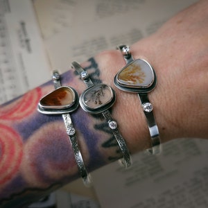 Puede incluir: Tres pulseras de plata con detalles de piedra de &aacute;gata. Cada pulsera presenta una piedra de &aacute;gata diferente con patrones y colores &uacute;nicos. Las pulseras est&aacute;n adornadas con peque&ntilde;as piedras preciosas transparentes.