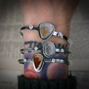 Puede incluir: Tres pulseras de plata con detalles de piedra de &aacute;gata y peque&ntilde;as piedras preciosas transparentes. Las pulseras est&aacute;n apiladas en la mu&ntilde;eca de una persona.