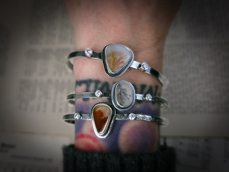 Puede incluir: Tres pulseras de plata con detalles de piedra de &aacute;gata y peque&ntilde;as piedras preciosas transparentes. Las pulseras est&aacute;n apiladas en la mu&ntilde;eca de una persona.