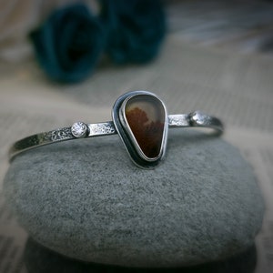 Puede incluir: Un brazalete de plata con una piedra de &aacute;gata en forma de l&aacute;grima engastada en un bisel de plata. La piedra tiene un patr&oacute;n marr&oacute;n y blanco. Hay dos peque&ntilde;os acentos plateados a cada lado de la piedra.