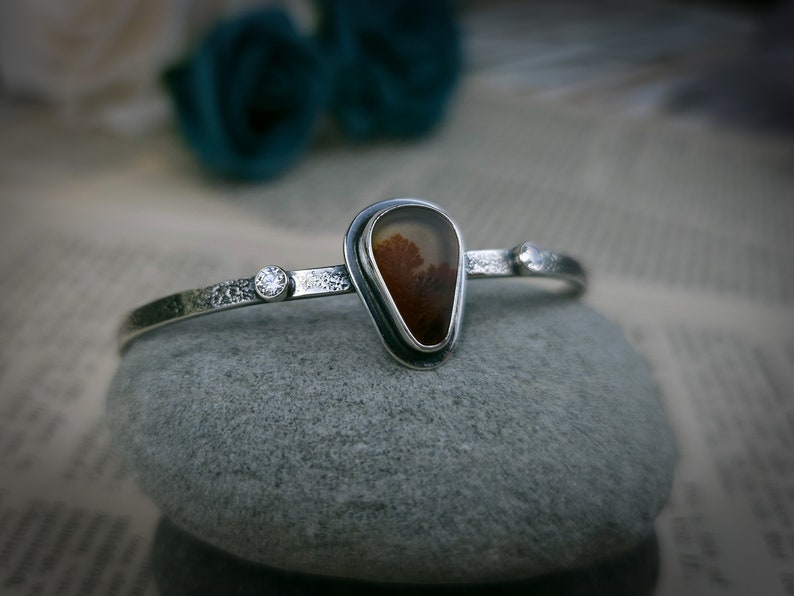 Puede incluir: Un brazalete de plata con una piedra de &aacute;gata en forma de l&aacute;grima engastada en un bisel de plata. La piedra tiene un patr&oacute;n marr&oacute;n y blanco. Hay dos peque&ntilde;os acentos plateados a cada lado de la piedra.
