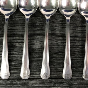 May include: Seven silver spoons arranged in a row on a dark wooden surface. The spoons have a classic design with oval bowls and long, slender handles. The spoons are reflective, and the wood grain is visible.