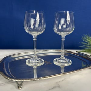 May include: Two clear wine glasses with long stems sit on a silver serving tray. The glasses are empty and reflect on the tray's surface. The tray has a beaded edge and decorative feet. A blue background is behind the tray.