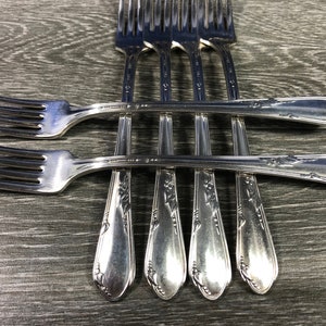 May include: A collection of silver forks arranged on a gray wooden surface. The forks have a classic design with tines and decorative handles. The arrangement showcases the silverware's elegant details, perfect for dining.