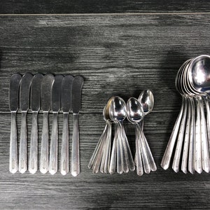 May include: A collection of vintage silverware, including a butter knife, butter spreaders, teaspoons, and soup spoons. The silverware is arranged on a dark gray wooden surface, showcasing the reflective quality of the polished metal.