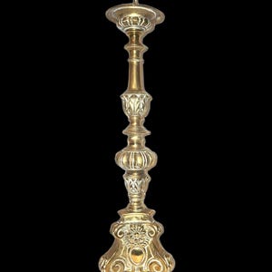 May include: An antique brass candlestick. It has a tall, ornate design with a wide base and a circular top for holding a candle. The candlestick features detailed carvings and a polished finish. The overall style is reminiscent of a classic, vintage aesthetic.