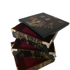 May include: A stack of four black lacquered boxes with gold and white bamboo and floral designs. The boxes have red velvet interiors and gold corner accents. The top box has a lid with a similar design.