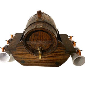 May include: A wooden beer barrel with a brass tap, mounted on a dark wood base. The barrel is secured with metal bands and has a wooden tap. Six ceramic beer steins are attached to the sides of the base.