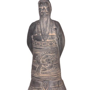 May include: A dark grey statue of a man in traditional Asian attire. The statue has a long beard and moustache, a tall hat, and intricate carvings of a dragon on the front of the robe. The figure's hands are clasped behind its back.
