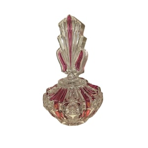 Könnte beinhalten: Eine klare Parfümflasche aus Glas mit einem rosa und klaren Verschluss. Die Flasche hat einen runden Boden mit geripptem Design und einen dekorativen, fächerförmigen Verschluss. Die Flasche ist wahrscheinlich Vintage und verleiht einen Hauch von Eleganz.