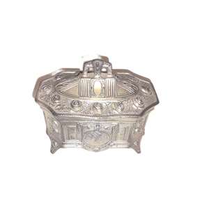 Könnte beinhalten: Silberfarbene, verzierte Schmuckschatulle mit Klappdeckel und dekorativen floralen und geometrischen Mustern. Die Schatulle hat kleine Füße und einen Griff oben. Das Gesamtdesign ist vintage und elegant.