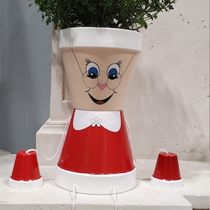 Peut inclure: Un pot de fleurs blanc avec un visage peint, portant une robe rouge et un col blanc, avec une fleur blanche sur le col. Le pot a des yeux bleus, des joues roses et un sourire noir. Le pot est posé sur un rebord blanc et a quatre pots rouges et blancs qui pendent de lui.