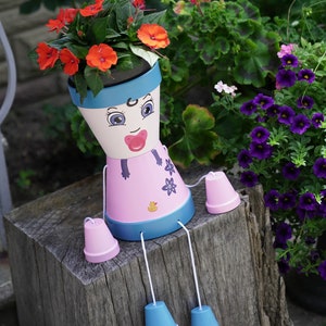 May include: A pink and blue painted flower pot shaped like a person with a painted face, wearing a pink dress and blue shoes. The pot is sitting on a wooden stump and has flowers growing out of the top.