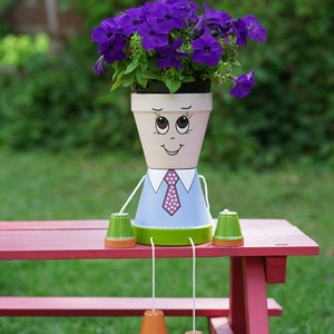 Peut inclure: Un pot de fleurs peint en forme de personne avec une chemise bleue, un pantalon vert et une cravate blanche à pois. Le pot contient des fleurs violettes et est posé sur une table de pique-nique rouge. Le texte "flowerpotpeople.ca" est visible en arrière-plan.