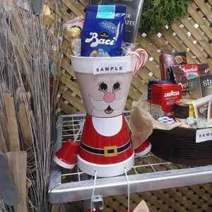 May include: A Santa Claus planter with a red and white hat, red and white beard, and a red belt. The planter is filled with a variety of gifts, including chocolate, candy canes, and a box of chocolates. The planter is labeled "SAMPLE".