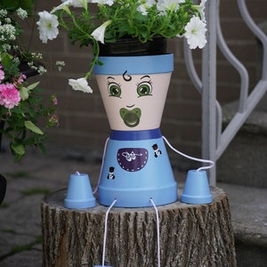 May include: A blue and pink painted flowerpot shaped like a baby with a dummy, wearing a blue overall with a white bib and brown bear designs. The pot has four blue legs and is sitting on a tree stump.
