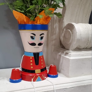 May include: A painted terracotta planter shaped like a nutcracker soldier with a red and blue uniform, holding a bouquet of orange and yellow chrysanthemums. The planter has four red and blue legs attached by strings.